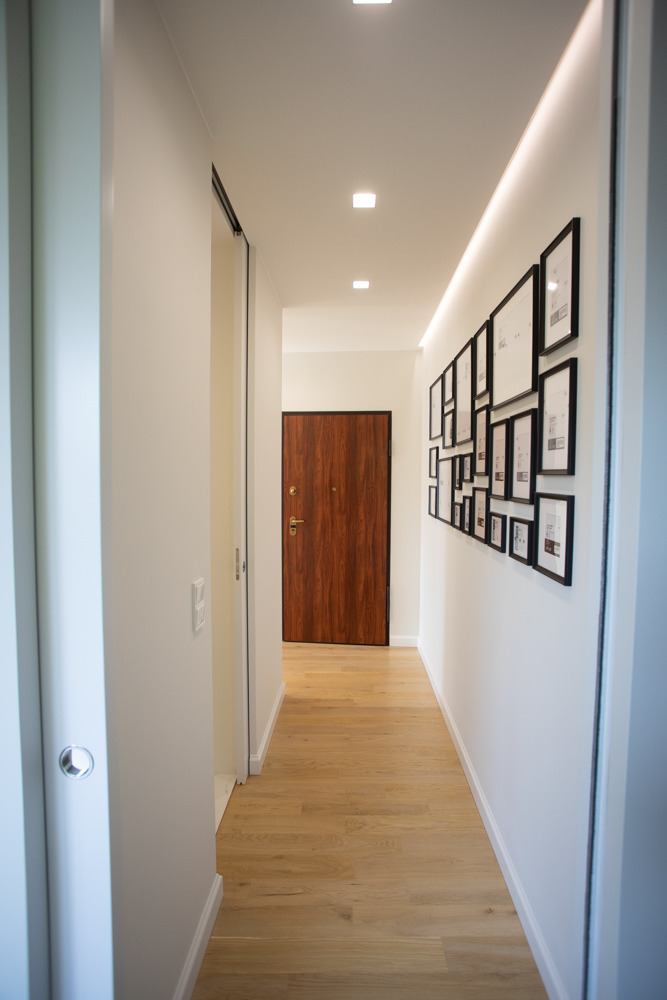 Nowoczesny korytarz z drewnianą podłogą, białymi ścianami, oświetleniem LED i galerią czarno-białych zdjęć w czarnych ramkach na ścianie.