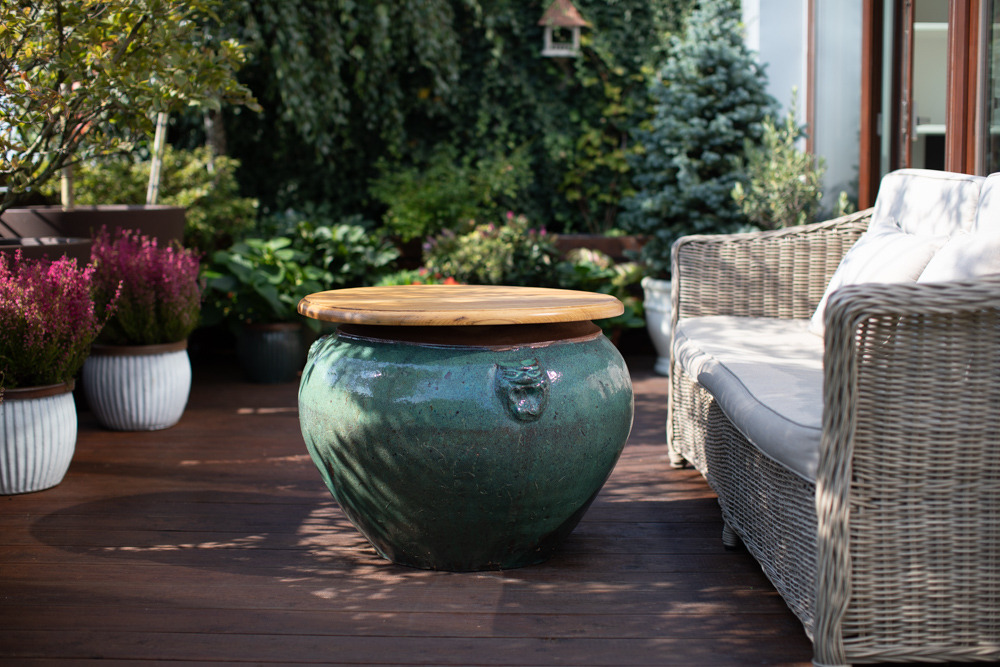 Zielony ceramiczny stolik ogrodowy z drewnianym blatem na drewnianym tarasie, obok wiklinowej sofy z poduszkami i roślin doniczkowych.