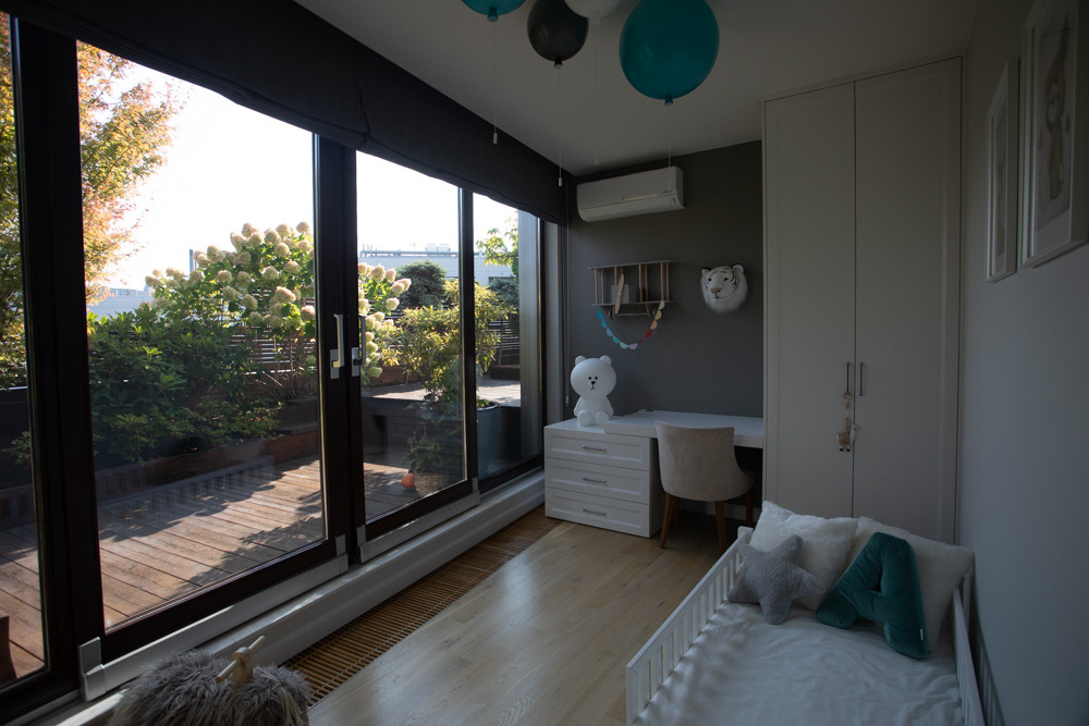 Jasny pokój dziecięcy z dużym oknem na taras, biurkiem z lampką w kształcie misia i łóżeczkiem z poduszkami w kształcie gwiazdy i litery A.