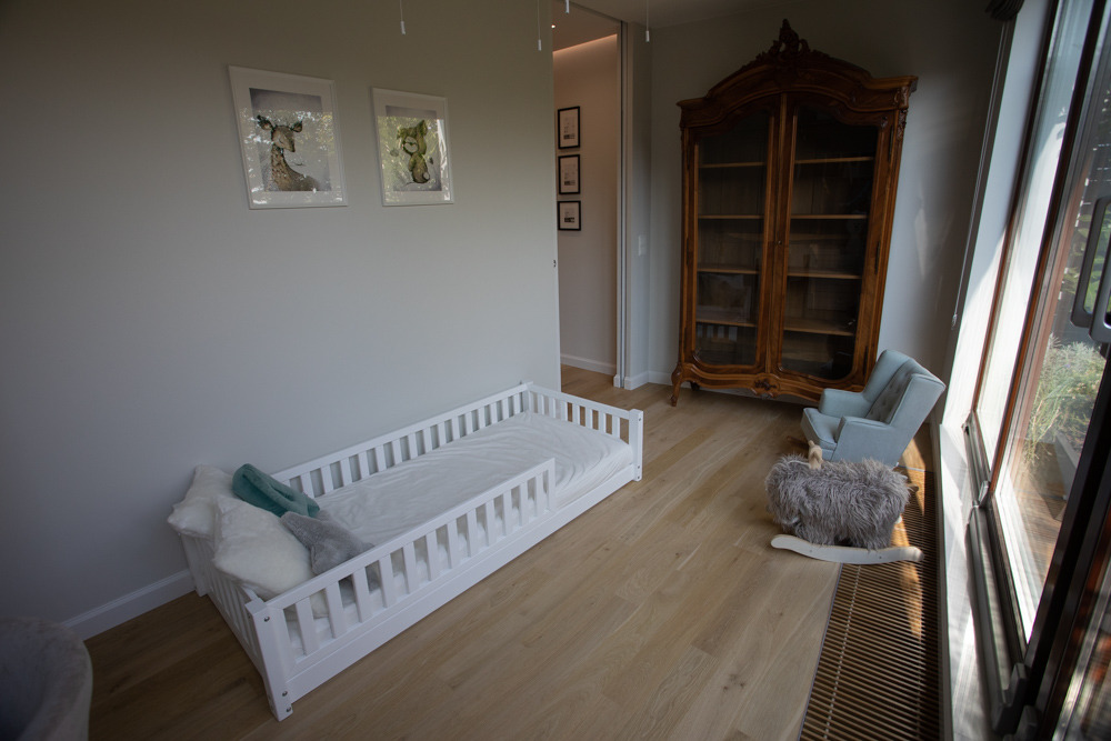 Jasne wnętrze pokoju dziecięcego z białym łóżeczkiem ze szczebelkami, drewnianą podłogą i antyczną witryną, oświetlone naturalnym światłem z okna.