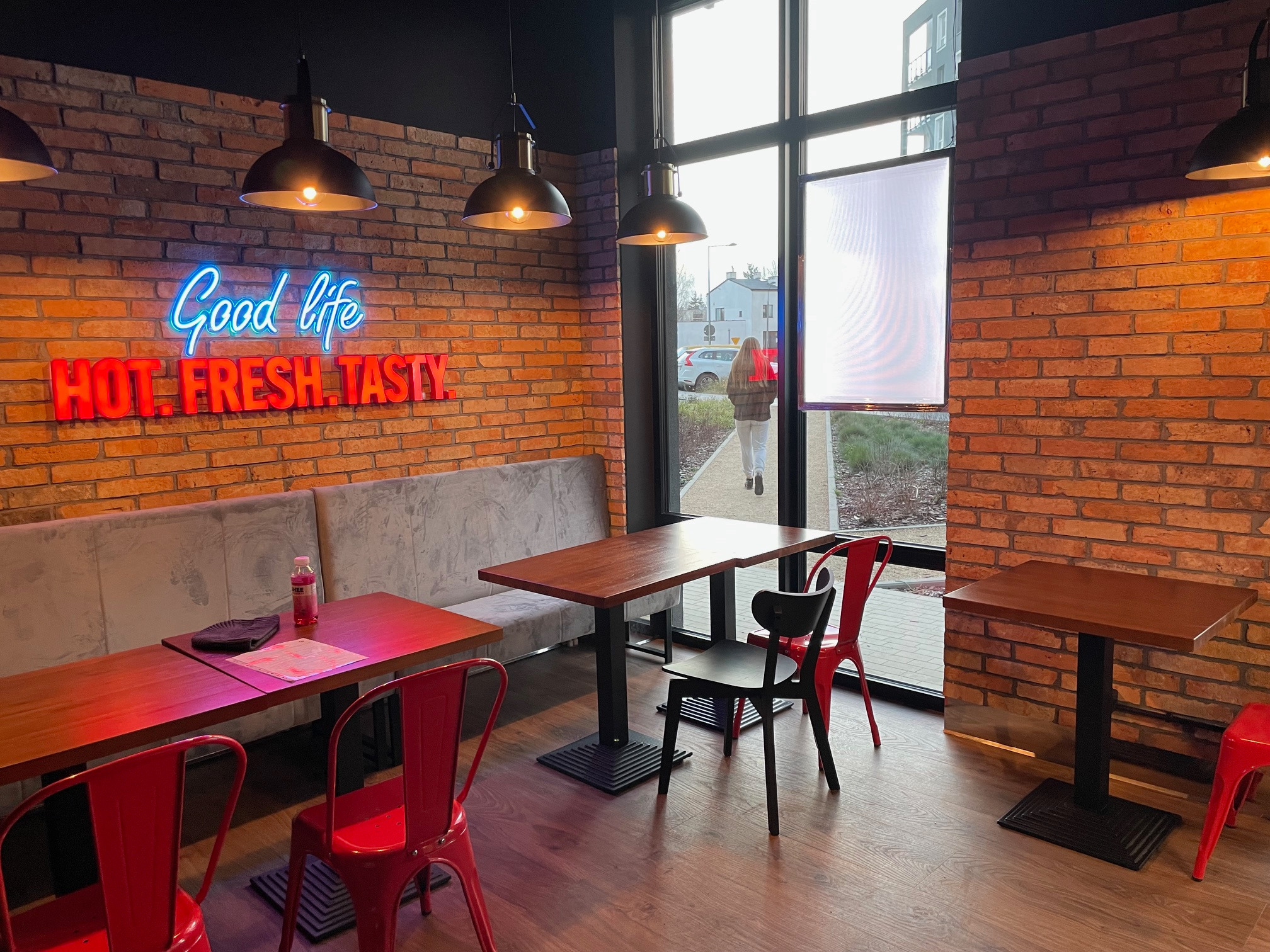 Wnętrze restauracji z ceglanymi ścianami, neonowym napisem 'Good life. Hot. Fresh. Tasty.', drewnianymi stołami, czerwonymi krzesłami i szarą sofą wzdłuż ściany.