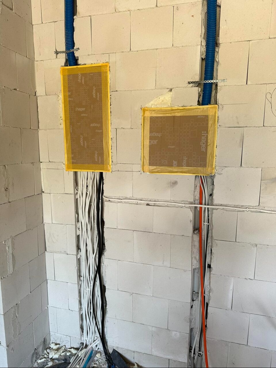 Wykonywanie bruzd w ścianie z betonu komórkowego pod przewody elektryczne, zabezpieczone kartonem marki Hager przyklejonym żółtą taśmą malarską, widoczne niebieskie rurki karbowane.