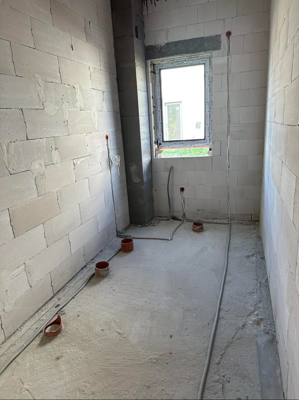 Surowy stan łazienki w bloku z widoczną instalacją elektryczną w peszlach na ścianach z betonu komórkowego i wylewce podłogowej, przygotowaną pod tynkowanie.