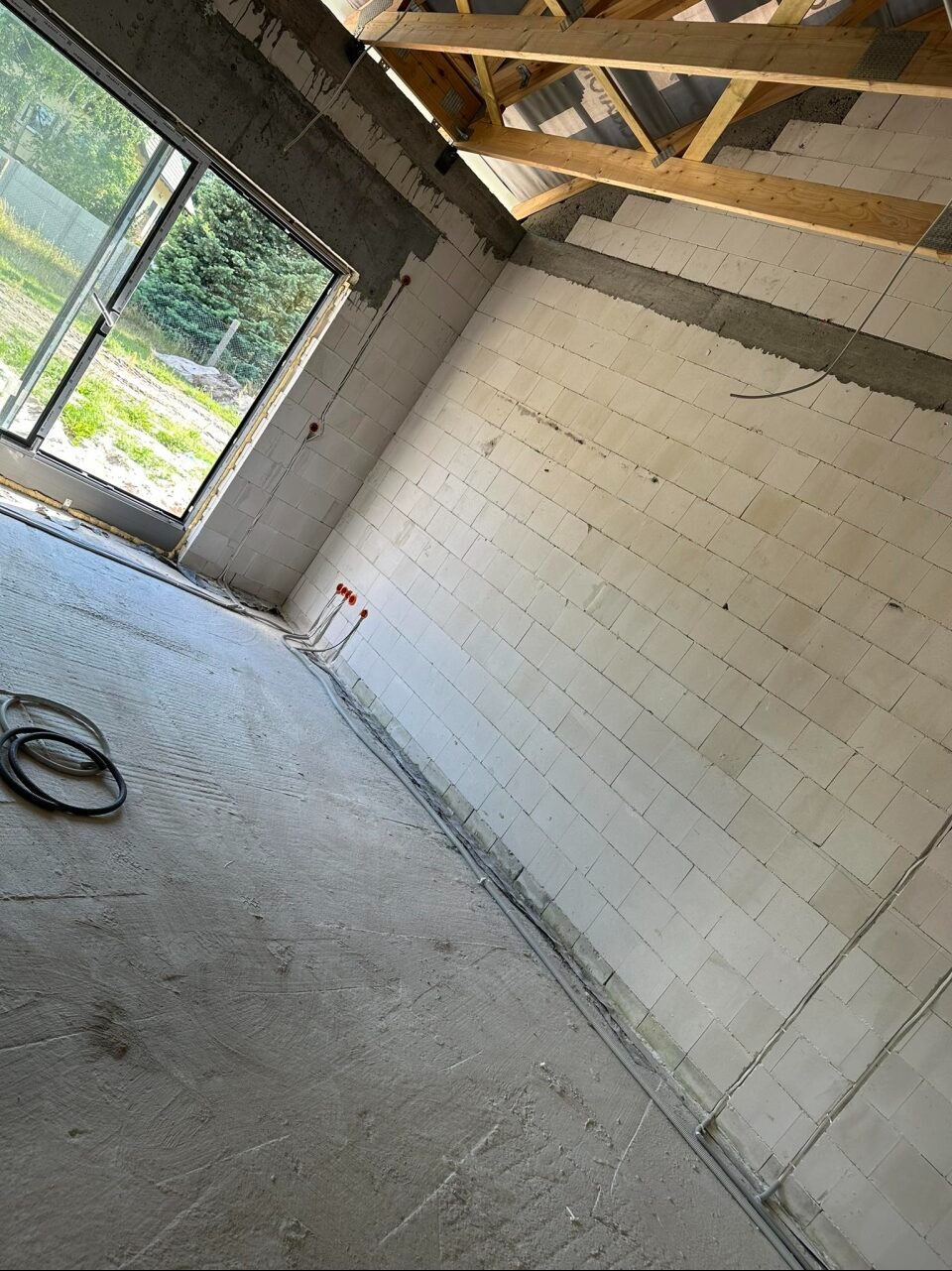 Surowy stan wnętrza budynku w trakcie budowy: ściany z białych bloczków, betonowa podłoga, widoczne przewody elektryczne wychodzące ze ściany, drewniana konstrukcja dachu, otwarte okno z widokiem...