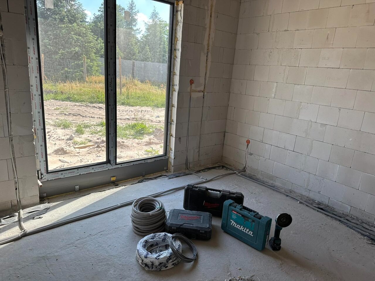 Wnętrze w stanie surowym w Nowej Soli z widocznymi przewodami elektrycznymi, puszkami, dwoma walizkami narzędziowymi Milwaukee i jedną Makita oraz szlifierką kątową akumulatorową Makita. Przewody...