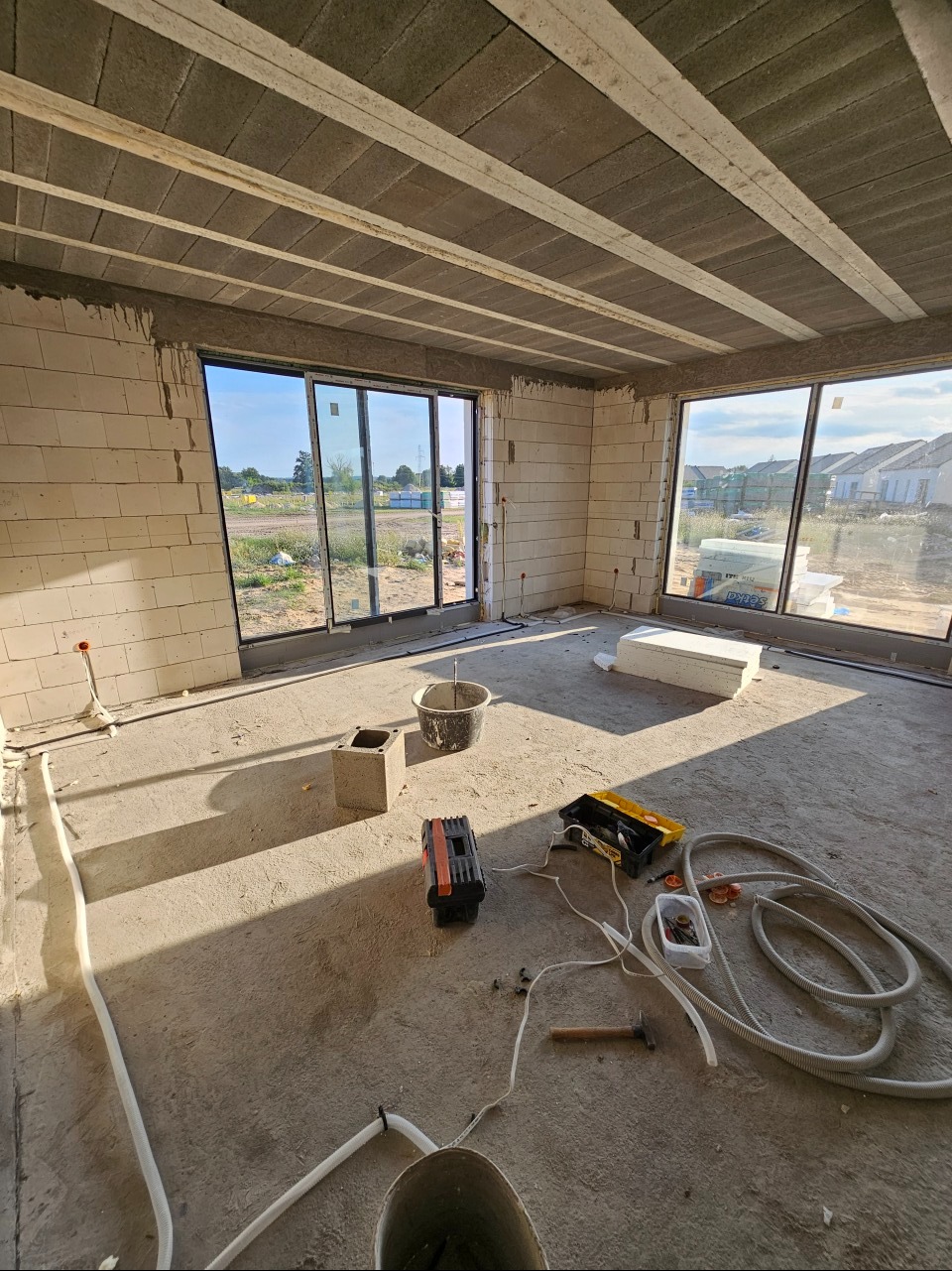 Wnętrze surowego domu w trakcie prac wykończeniowych, widoczne poprowadzone rury instalacyjne na podłodze, narzędzia i materiały budowlane, duże okna bez parapetów, ściany z białego pustaka.