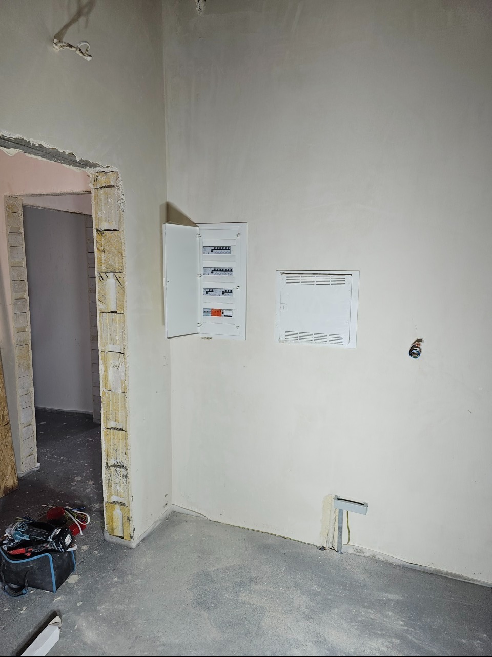 Otwarta skrzynka z bezpiecznikami na ścianie w surowym wnętrzu, widoczne kable i narzędzia w torbie na podłodze, ściany w trakcie wykańczania.