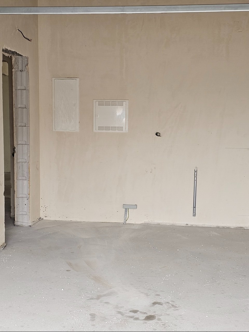 Surowe wnętrze w trakcie wykańczania: widoczne puszki instalacyjne na ścianie, surowy tynk, wylewka podłogowa z drobnymi zabrudzeniami, metalowa listwa pod sufitem.