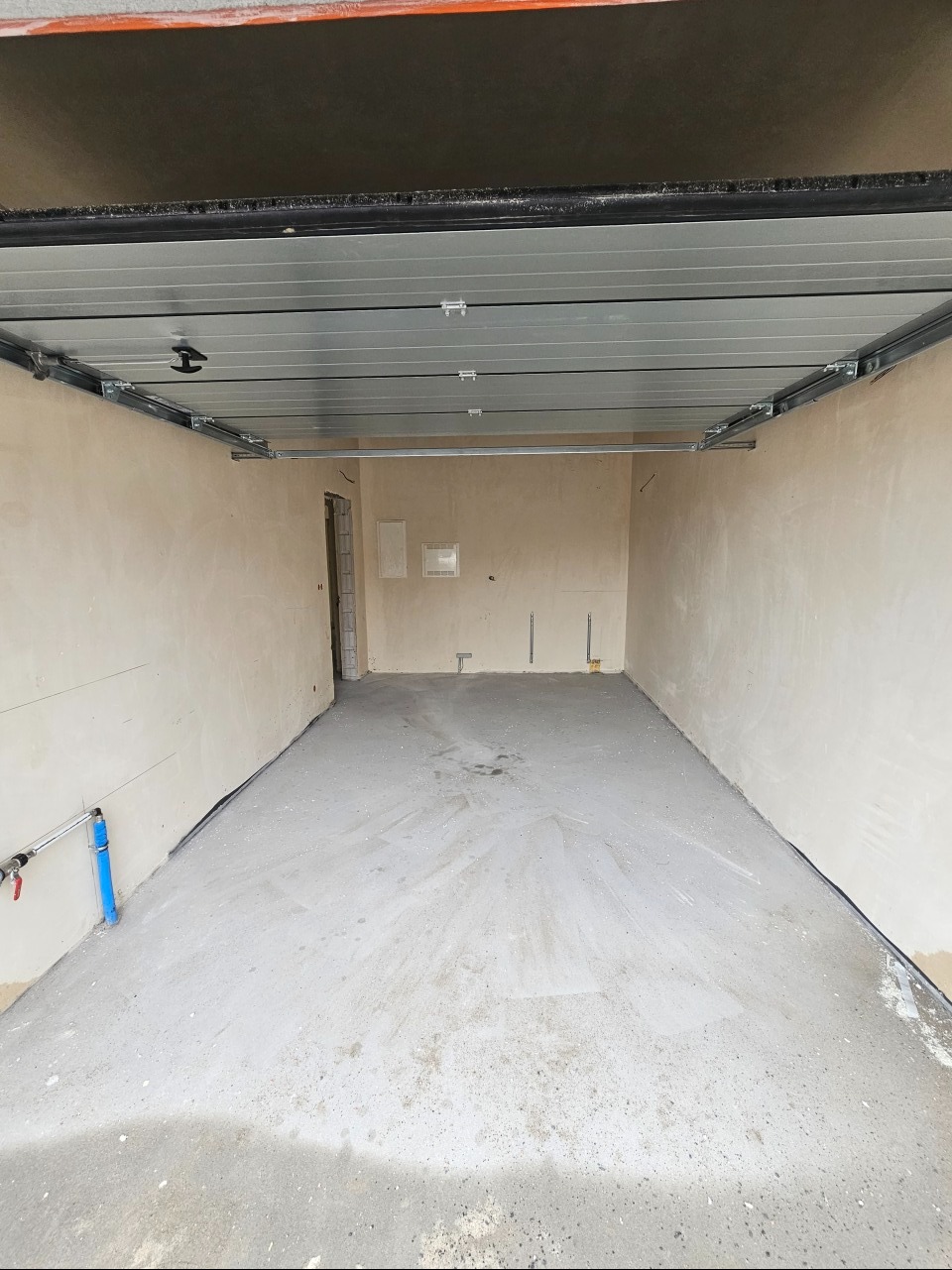 Wnętrze garażu w stanie surowym, widoczne metalowe szyny sufitowe do bramy segmentowej, ściany otynkowane, posadzka betonowa, instalacja wodna przy ścianie.
