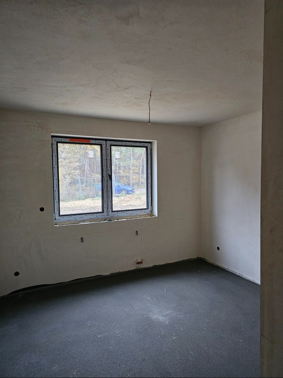 Surowe wnętrze z oknem, widoczne przewody elektryczne wystające ze ściany, szare wylewki podłogowe, ściany w trakcie tynkowania.