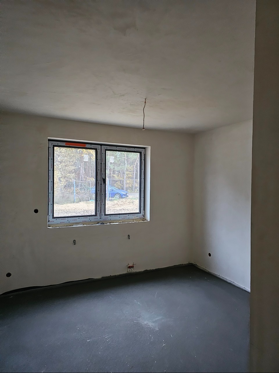 Surowe wnętrze pokoju w trakcie remontu: wylewka na podłodze, ściany po gładzi, okno PCV z widokiem na zewnątrz, wystający kabel elektryczny ze ściany.