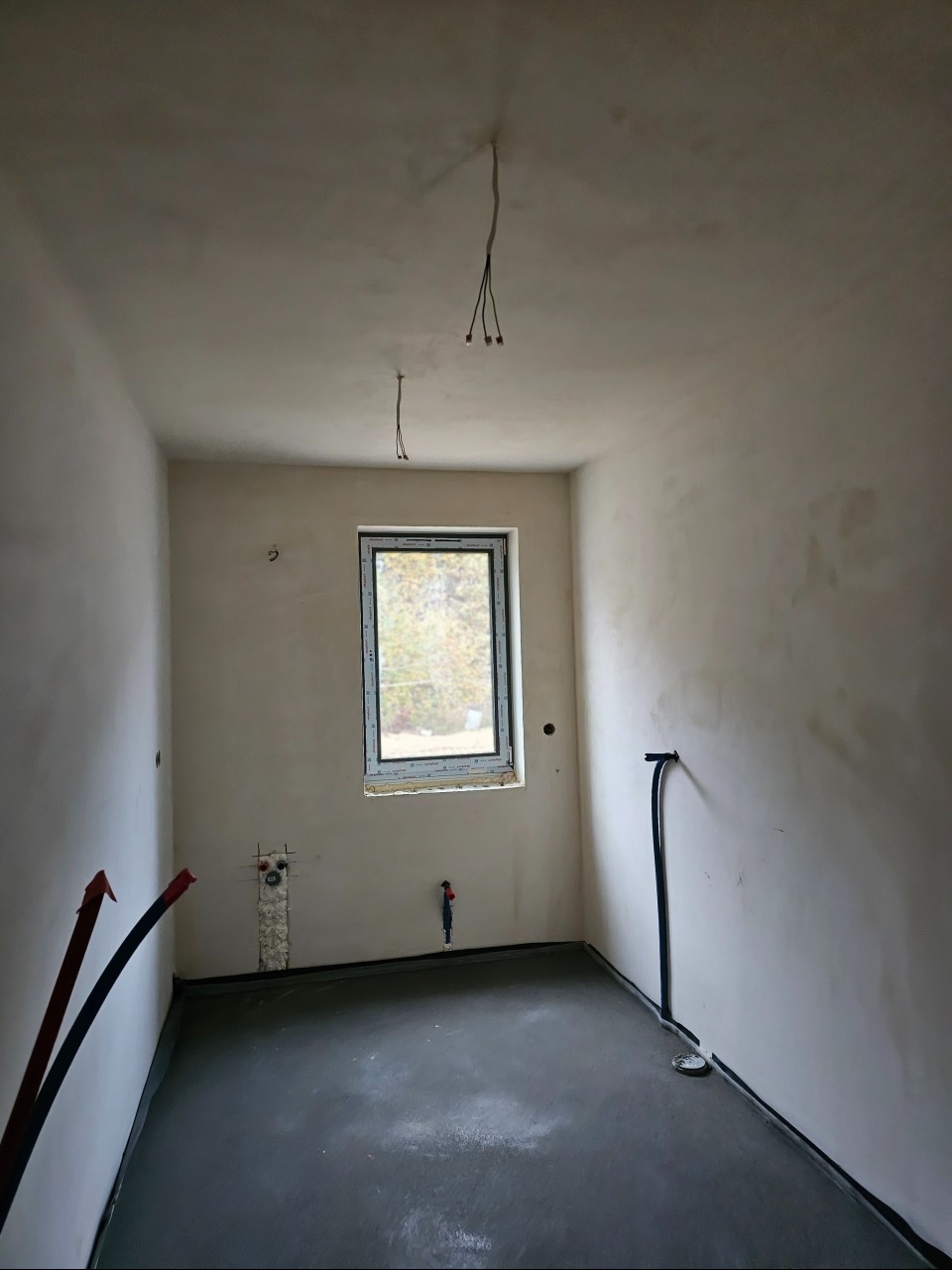 Surowy stan wnętrza łazienki z wyprowadzonymi przewodami elektrycznymi i hydraulicznymi, widoczne okno z widokiem na zewnątrz, szara wylewka na podłodze, ściany przygotowane do malowania.