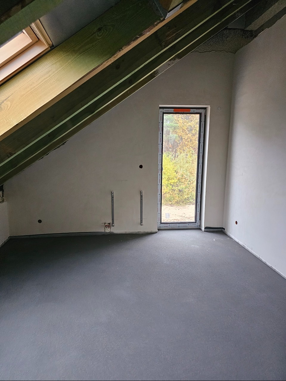 Poddasze w stanie deweloperskim z oknem i widocznymi elementami instalacji elektrycznej w ścianie.