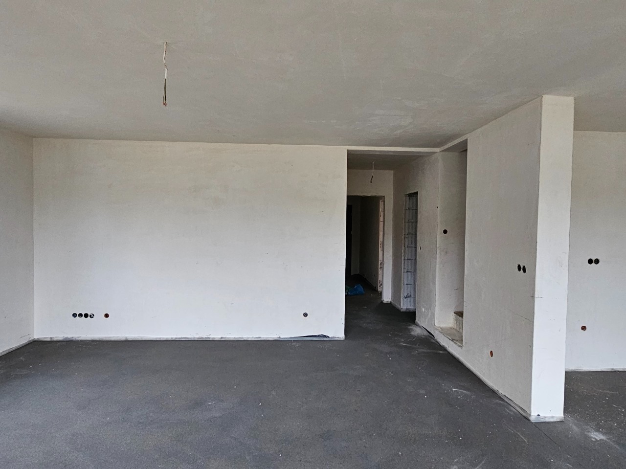 Surowe wnętrze w trakcie remontu: szare wylewki podłogowe, otynkowane białe ściany z widocznymi puszkami elektrycznymi i wystającymi przewodami z sufitu. Widoczny otwarty plan pomieszczeń...