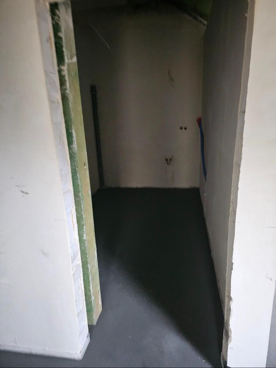 Surowe wnętrze łazienki w trakcie remontu: wylewka podłogowa, wystające kable i rury instalacji wodnej na ścianie, widoczne ościeżnice drzwi.