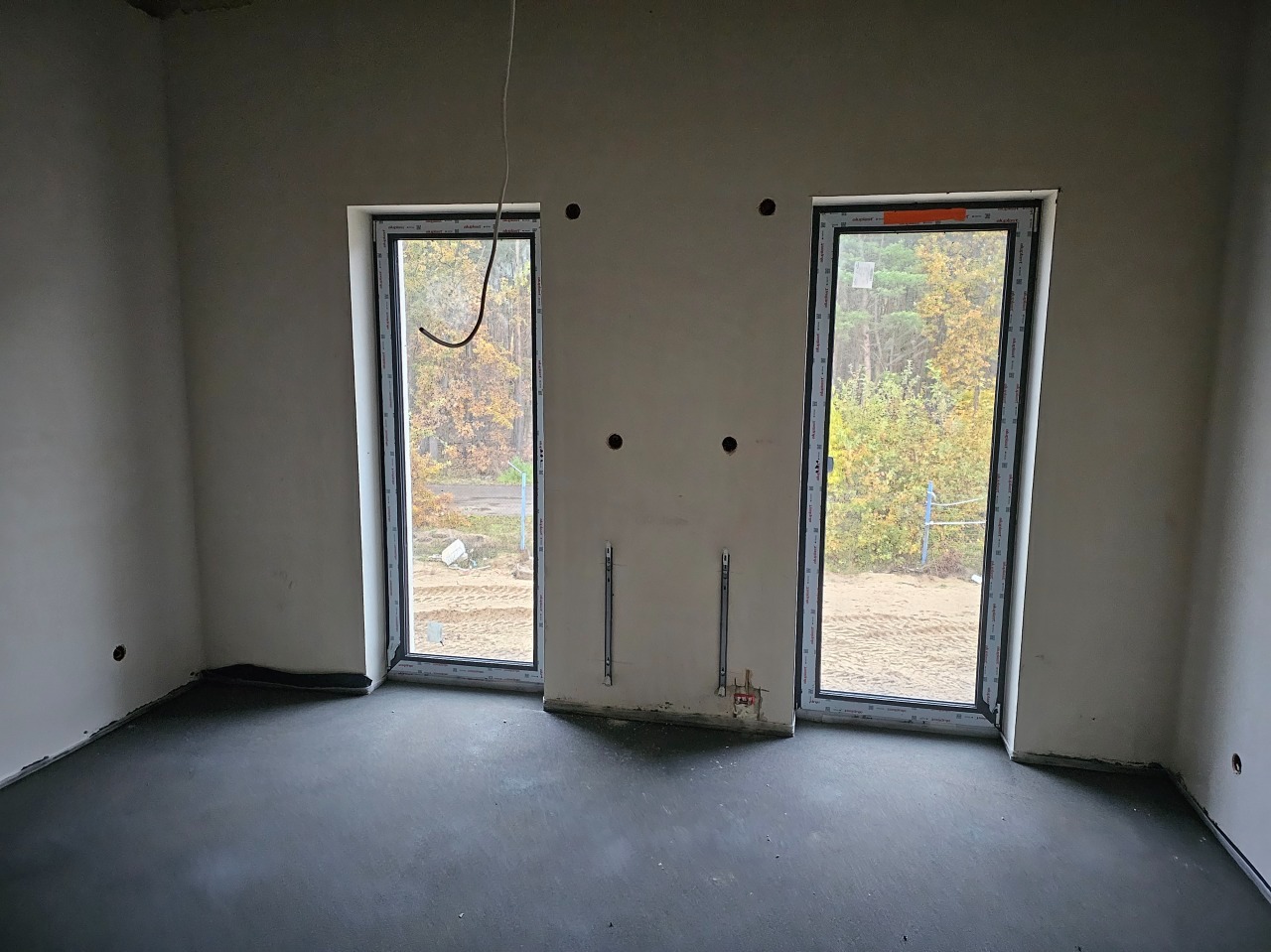Surowe wnętrze w trakcie wykańczania: dwa okna z widokiem na las, betonowa wylewka na podłodze, przygotowane punkty elektryczne na ścianach i wystający przewód elektryczny z sufitu.