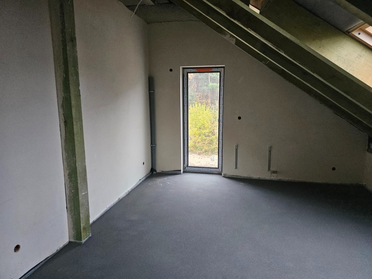 Surowe wnętrze poddasza z oknem dachowym i widoczną konstrukcją dachu, przygotowane do prac wykończeniowych, z szarą wylewką na podłodze i widocznymi elementami instalacji na ścianach.