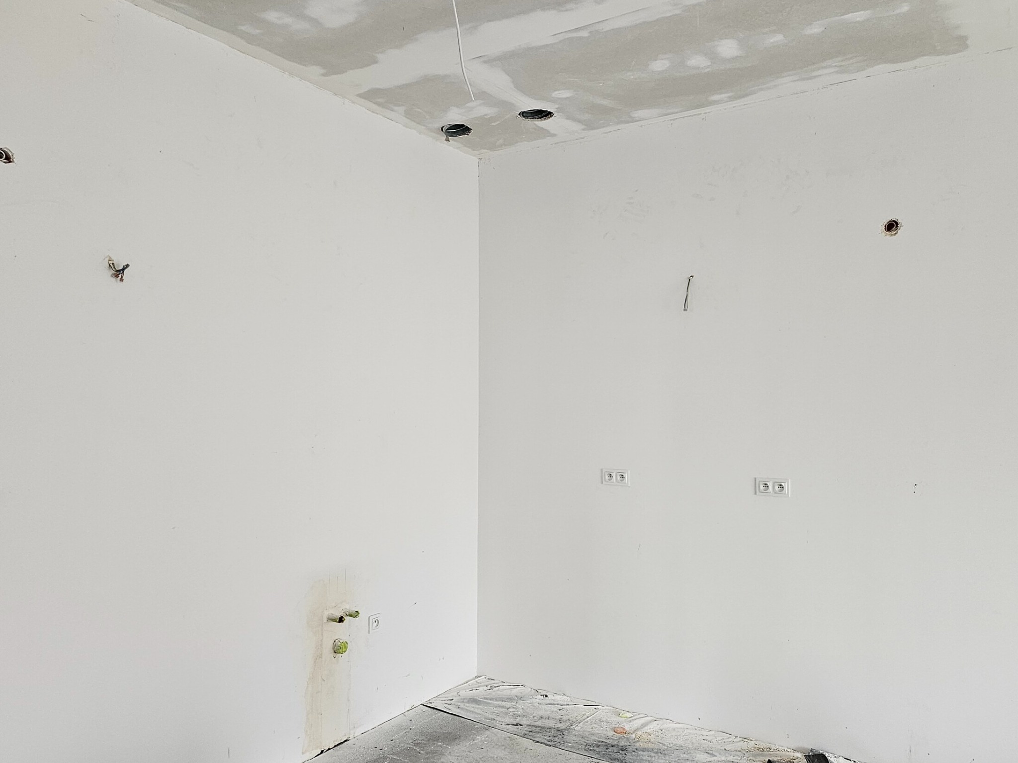 Surowe wnętrze w trakcie remontu: białe ściany z widocznymi przewodami elektrycznymi, gniazdkami i otworami w suficie, częściowo zagruntowany sufit, zabezpieczona podłoga.