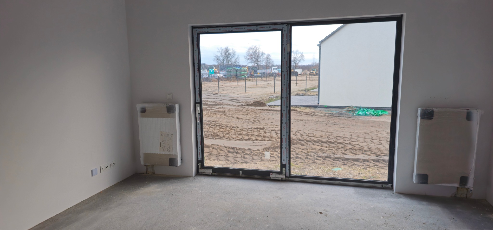 Wnętrze surowego domu z zamontowanymi, ale jeszcze ofoliowanymi grzejnikami na ścianach, widok na plac budowy przez duże przesuwne okno.
