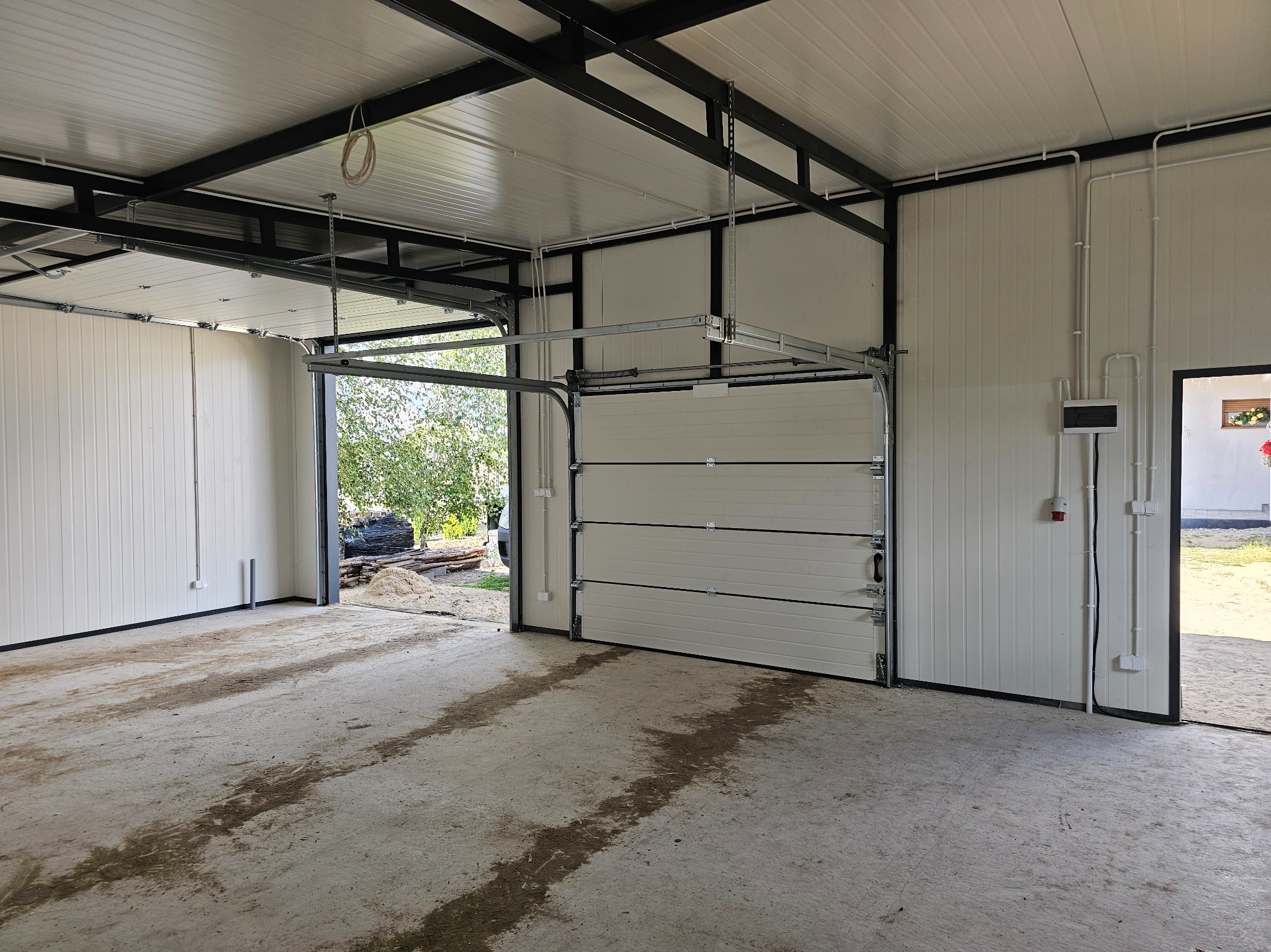 Wnętrze garażu z częściowo otwartą segmentową bramą garażową w kolorze złamanej bieli, widoczne instalacje elektryczne na ścianie i konstrukcja nośna dachu