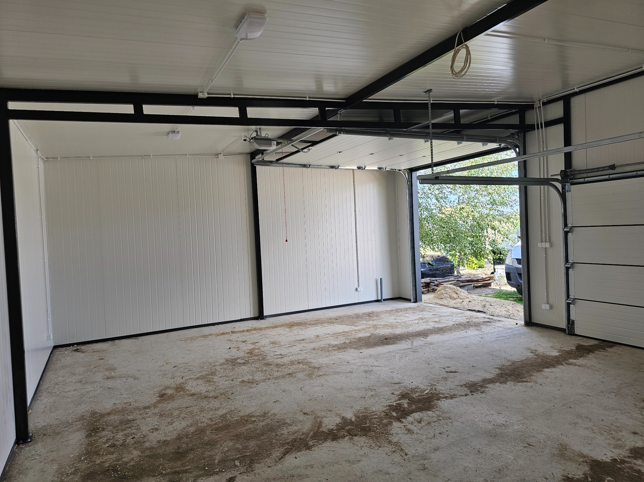 Wnętrze garażu z otwartą segmentową bramą garażową, widoczny napęd elektryczny na suficie, białe ściany z pionowymi panelami, betonowa posadzka z zabrudzeniami, na zewnątrz widoczne drzewa...