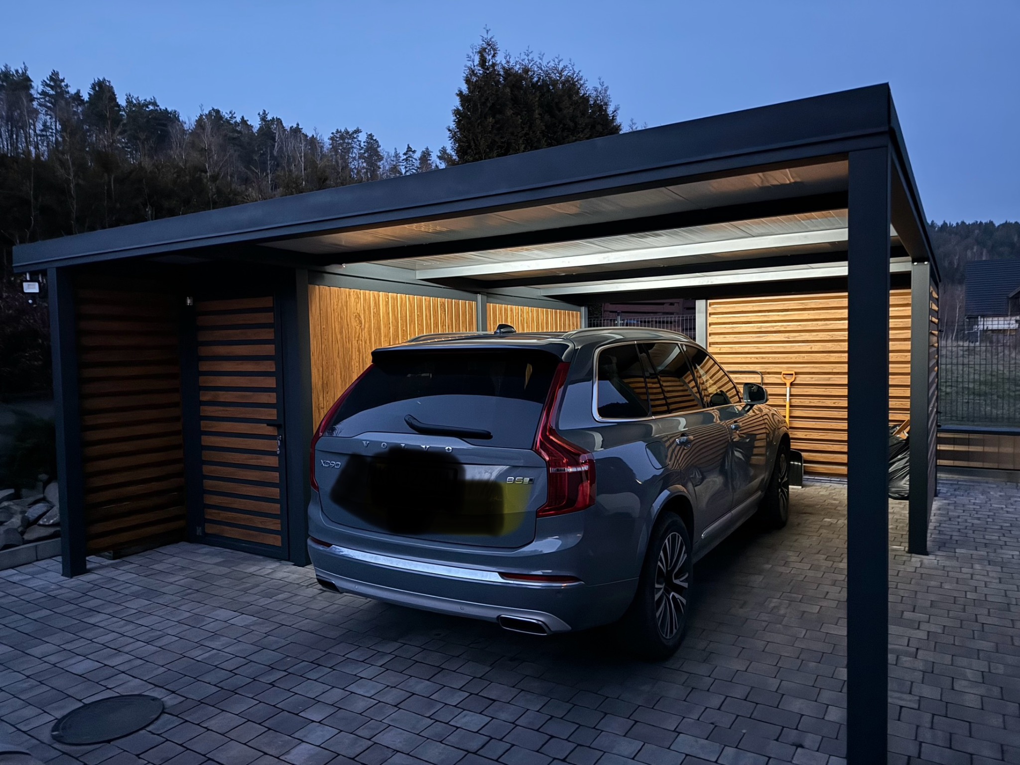 Nowoczesna, grafitowa wiata garażowa z drewnianymi panelami, oświetleniem LED i szarym SUV-em Volvo XC90 na kostce brukowej.