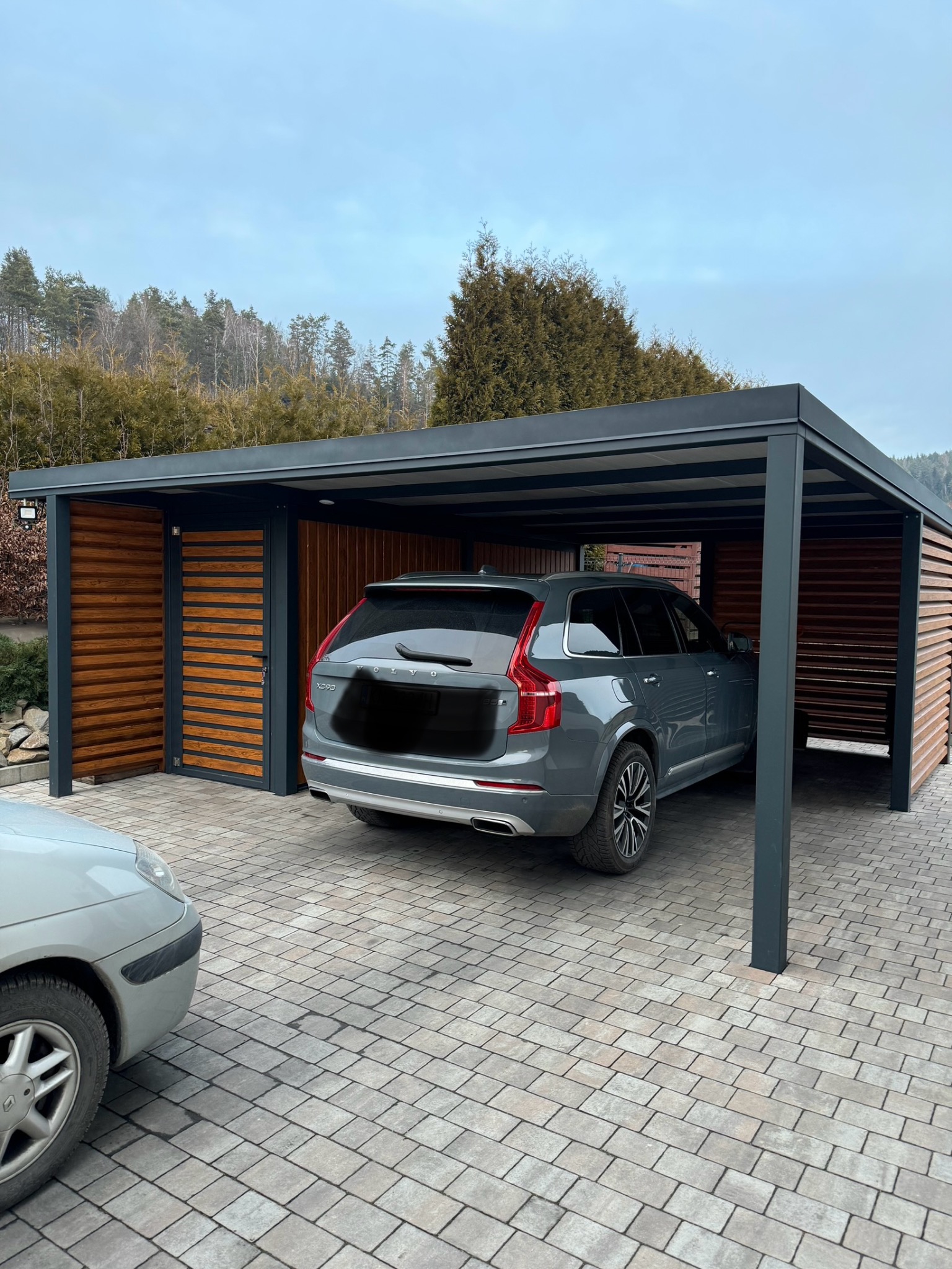 Szary samochód Volvo XC90 zaparkowany pod nowoczesną, zadaszoną konstrukcją z metalu i drewnianych paneli, z kostki brukowej podłoże i drzewa w tle.