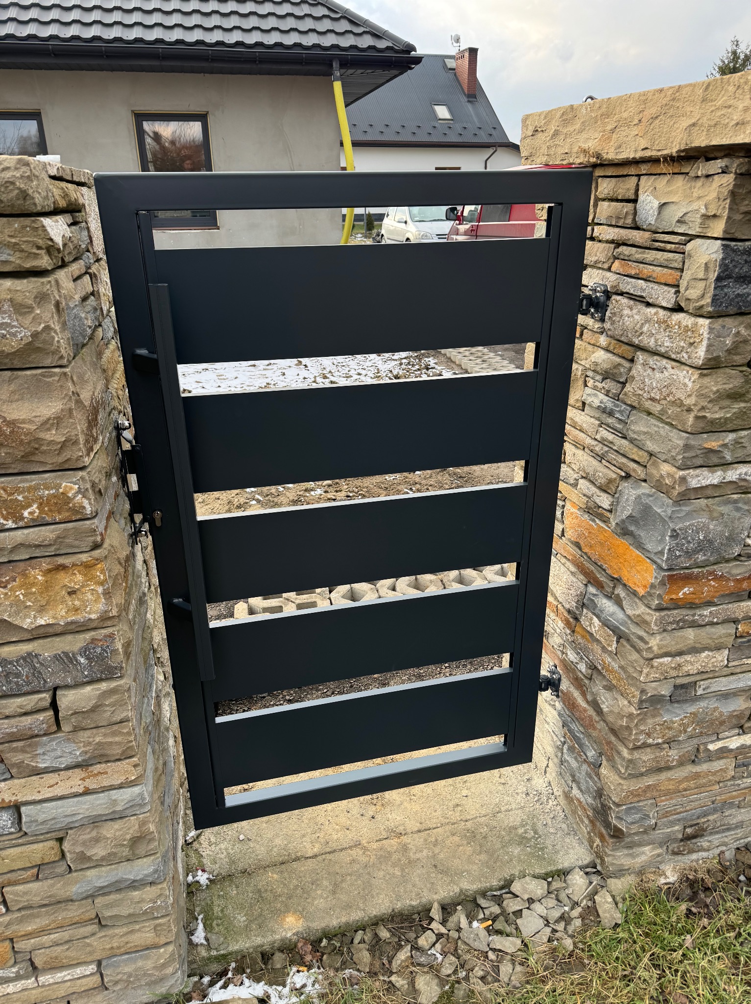 Nowoczesna, czarna brama z poziomymi szczeblami, osadzona w kamiennych słupkach z naturalnego kamienia łupanego w odcieniach beżu i szarości, widok na posesję z elementami kostki brukowej...