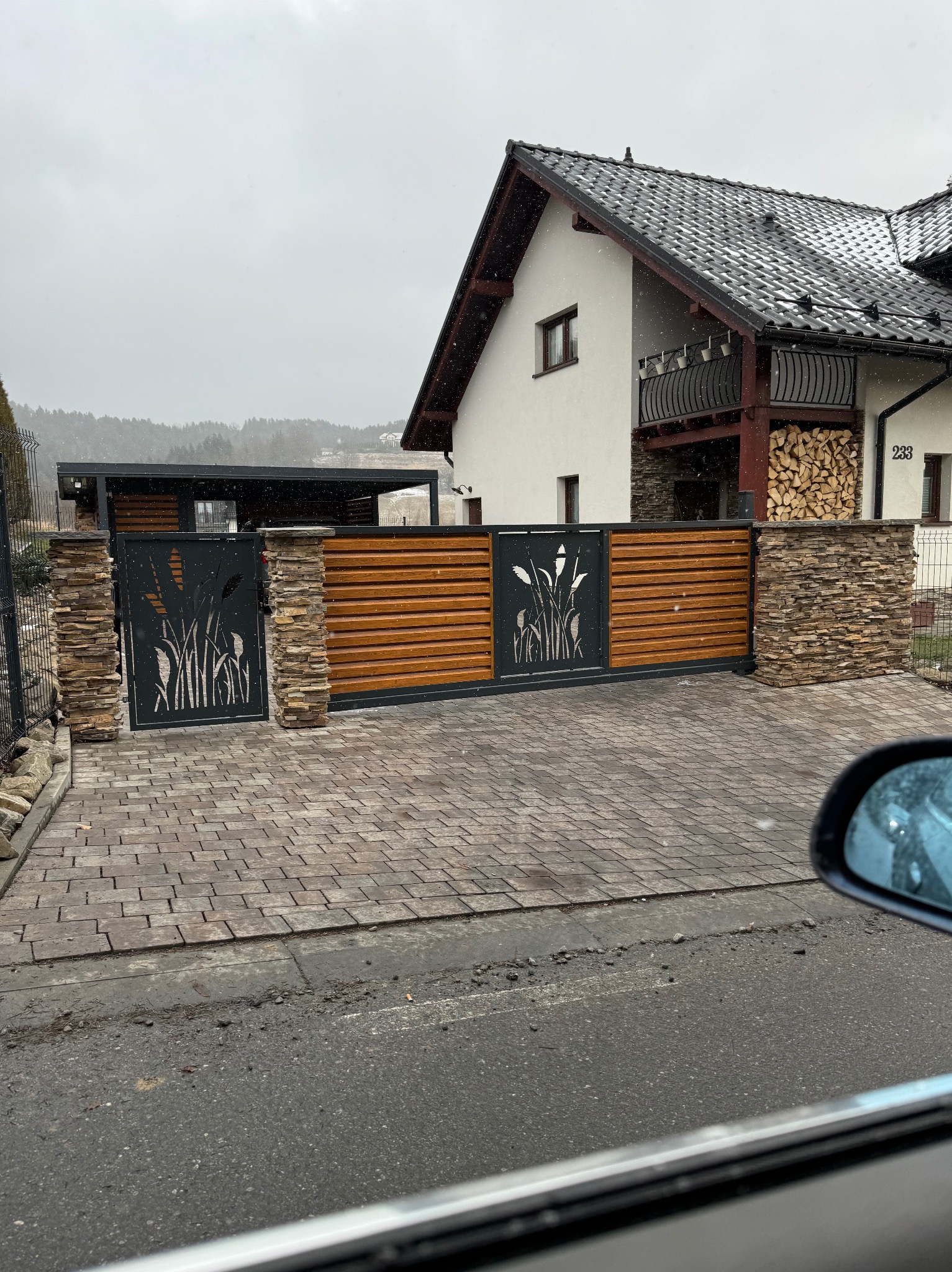 Nowoczesna brama wjazdowa z dekoracyjnym motywem roślinnym, połączona z drewnianym ogrodzeniem i kamiennymi słupkami, prowadząca do domu w zimowej scenerii.
