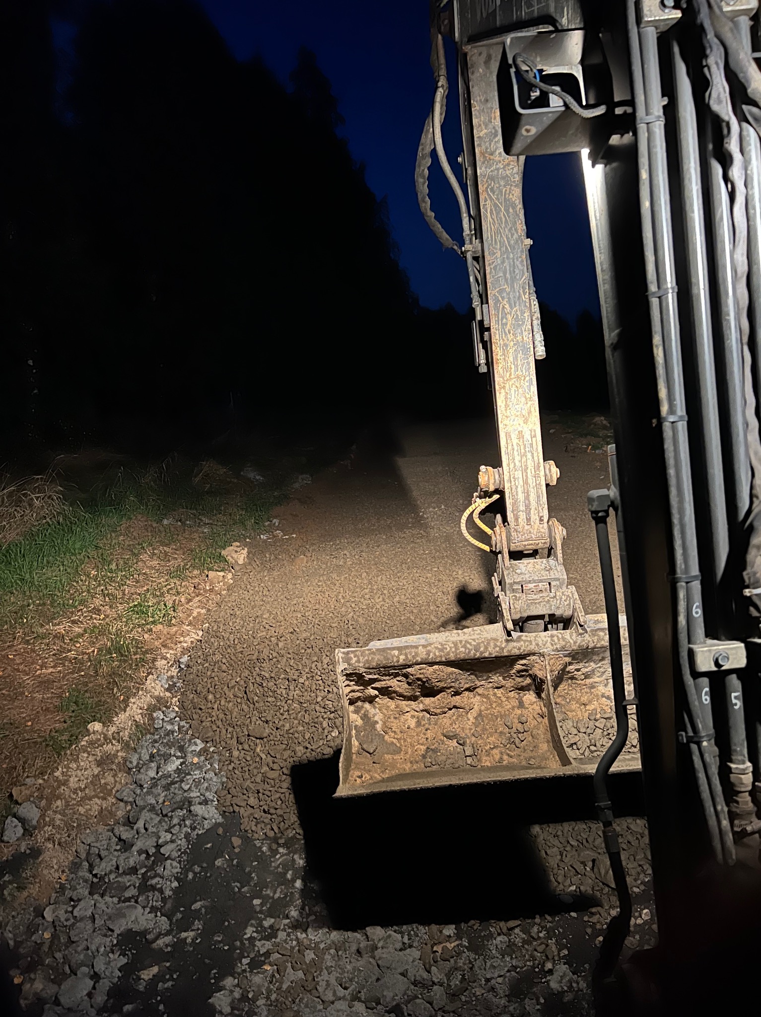 Koparka w trakcie niwelacji terenu pod osłoną nocy, oświetlona reflektorem, z widocznymi przewodami hydraulicznymi.