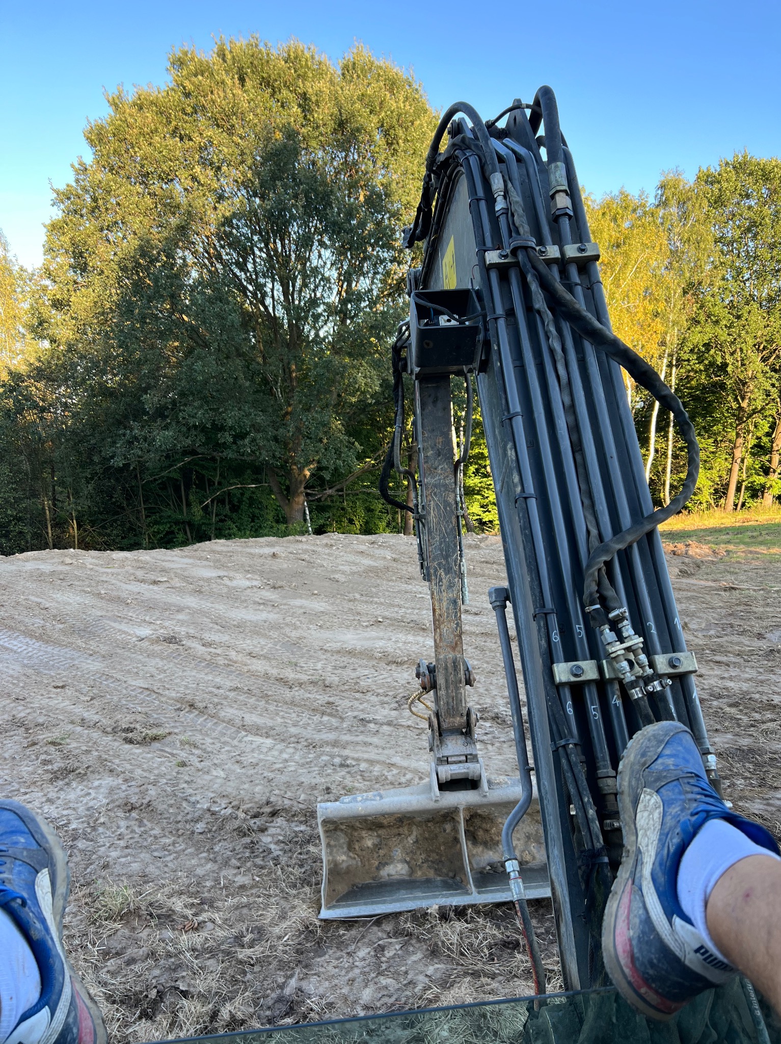 Widok z kabiny koparki na hałdę piasku i drzewa w tle; stopa w niebieskim bucie Puma oparta o ramię koparki.