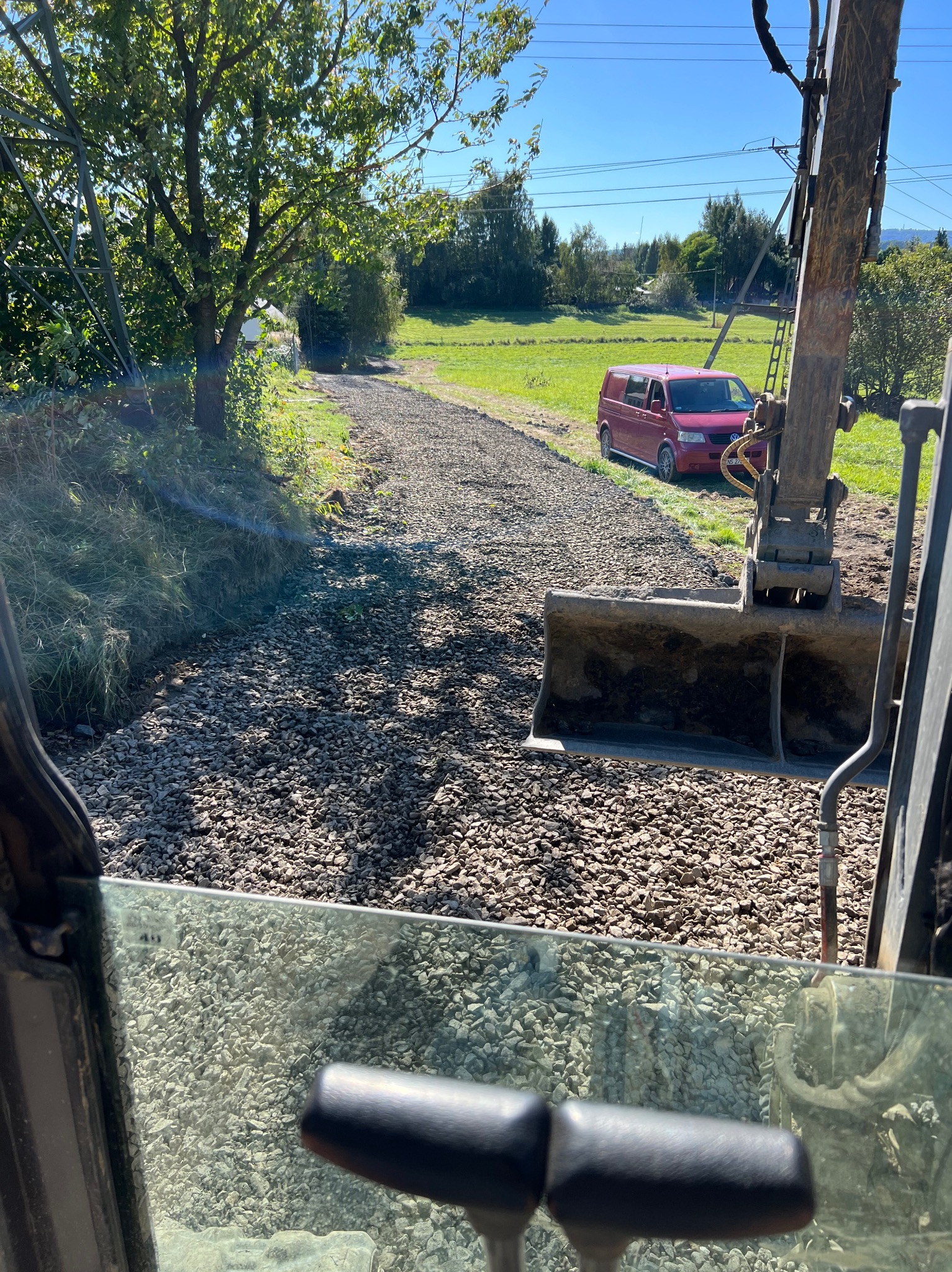 Widok z kabiny koparki na utwardzoną żwirem drogę, z zaparkowanym czerwonym vanem w tle i zielonym krajobrazem.