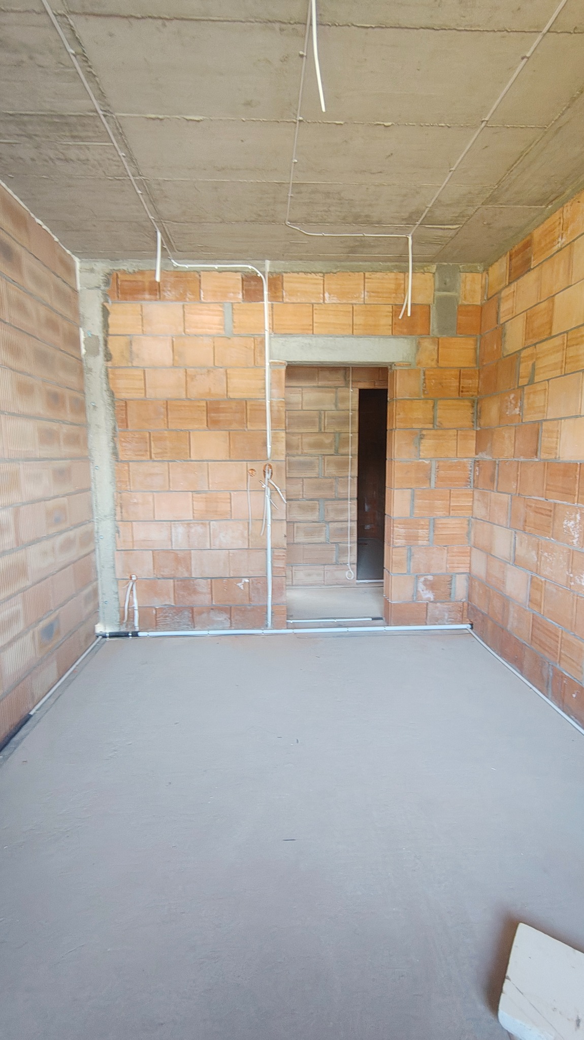 Surowy pokój z ceglanymi ścianami, betonową podłogą i sufitem. Widoczne białe rurki instalacji elektrycznej na ścianach i suficie. W tle otwór drzwiowy.