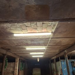 Robo Elektroinstalacje - Oświetlony korytarz w starym budynku gospodarczym z rzędami podłużnych lamp LED zamontowanych na betonowym suficie, z widocznymi przewodami instalacyjnymi i metalowymi belkami.