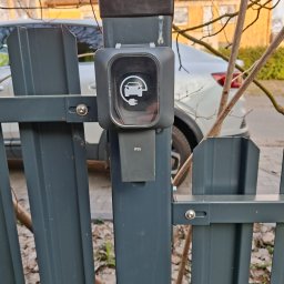 Robo Elektroinstalacje - Szara stacja ładowania samochodów elektrycznych z ikoną auta i wtyczki, o klasie szczelności IP55, zamontowana na ciemnoszarym metalowym ogrodzeniu, z białym autem w tle.