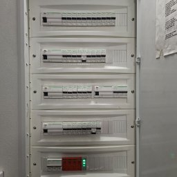 Robo Elektroinstalacje - Otwarta biała rozdzielnica elektryczna z pięcioma rzędami wyłączników nadprądowych Schneider Electric, z zielonymi diodami na dole, żółtą naklejką ostrzegawczą i listą obwodów na drzwiach.