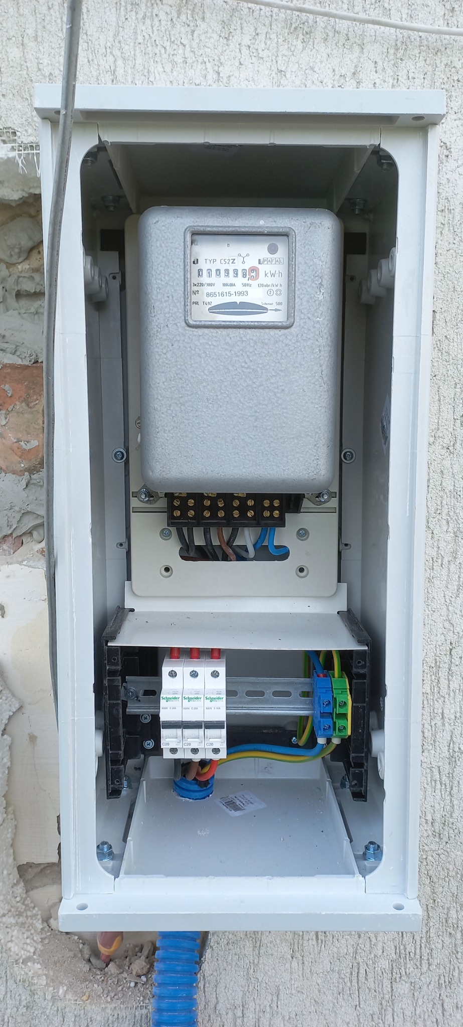 Szczegółowe ujęcie wnętrza skrzynki elektrycznej z zamontowanym licznikiem energii, bezpiecznikami i okablowaniem, na tle zewnętrznej ściany budynku.