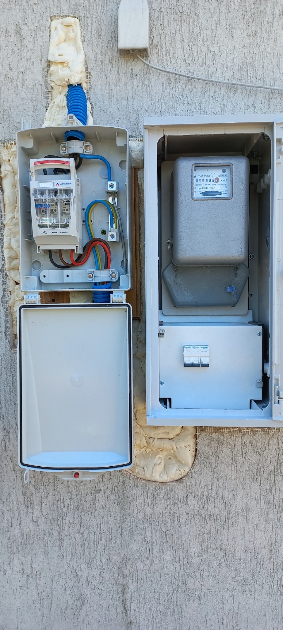 Otwarta skrzynka elektryczna z bezpiecznikami i licznikiem, zabezpieczona pianką montażową na elewacji budynku.