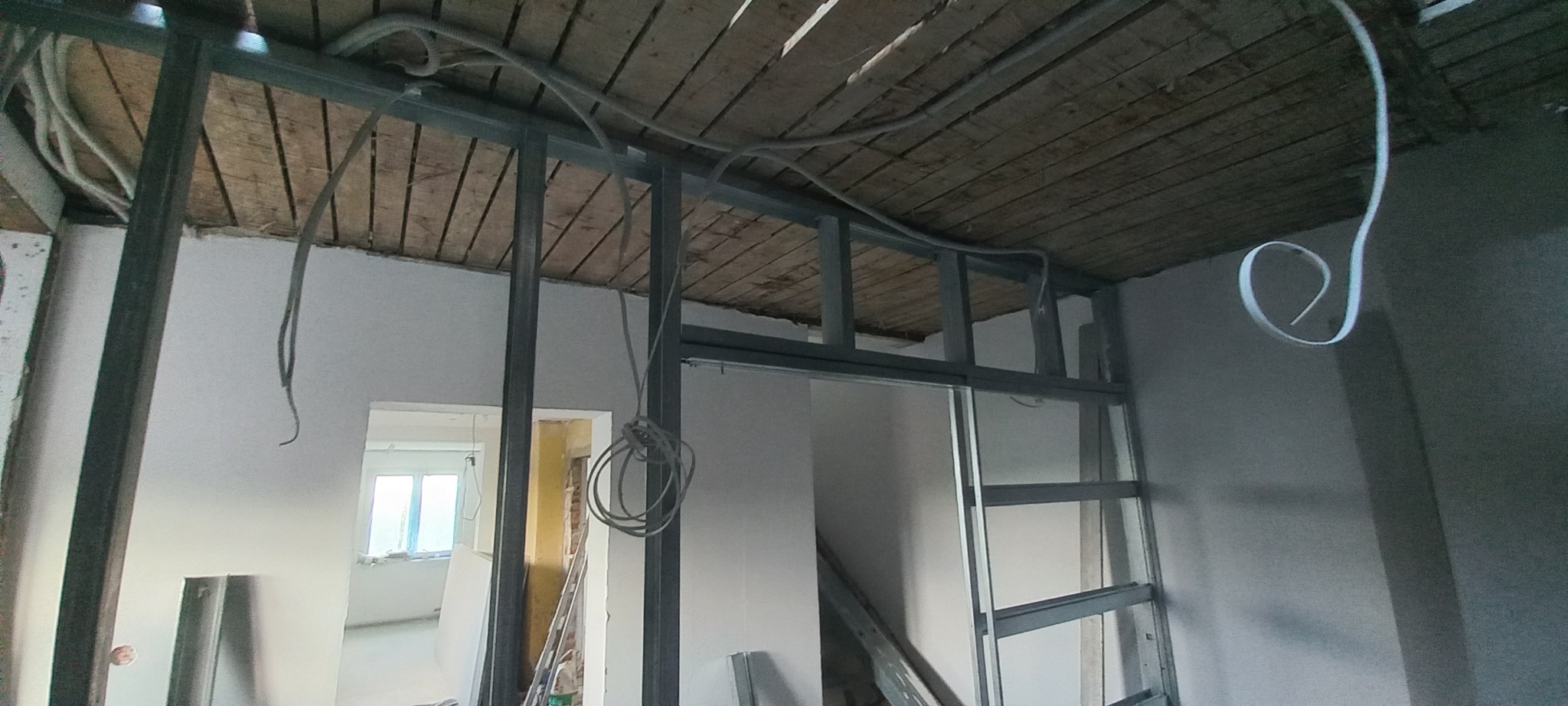 Wnętrze w trakcie remontu: metalowa konstrukcja szkieletowa ścian działowych, widoczne przewody elektryczne zwisające z sufitu z desek.