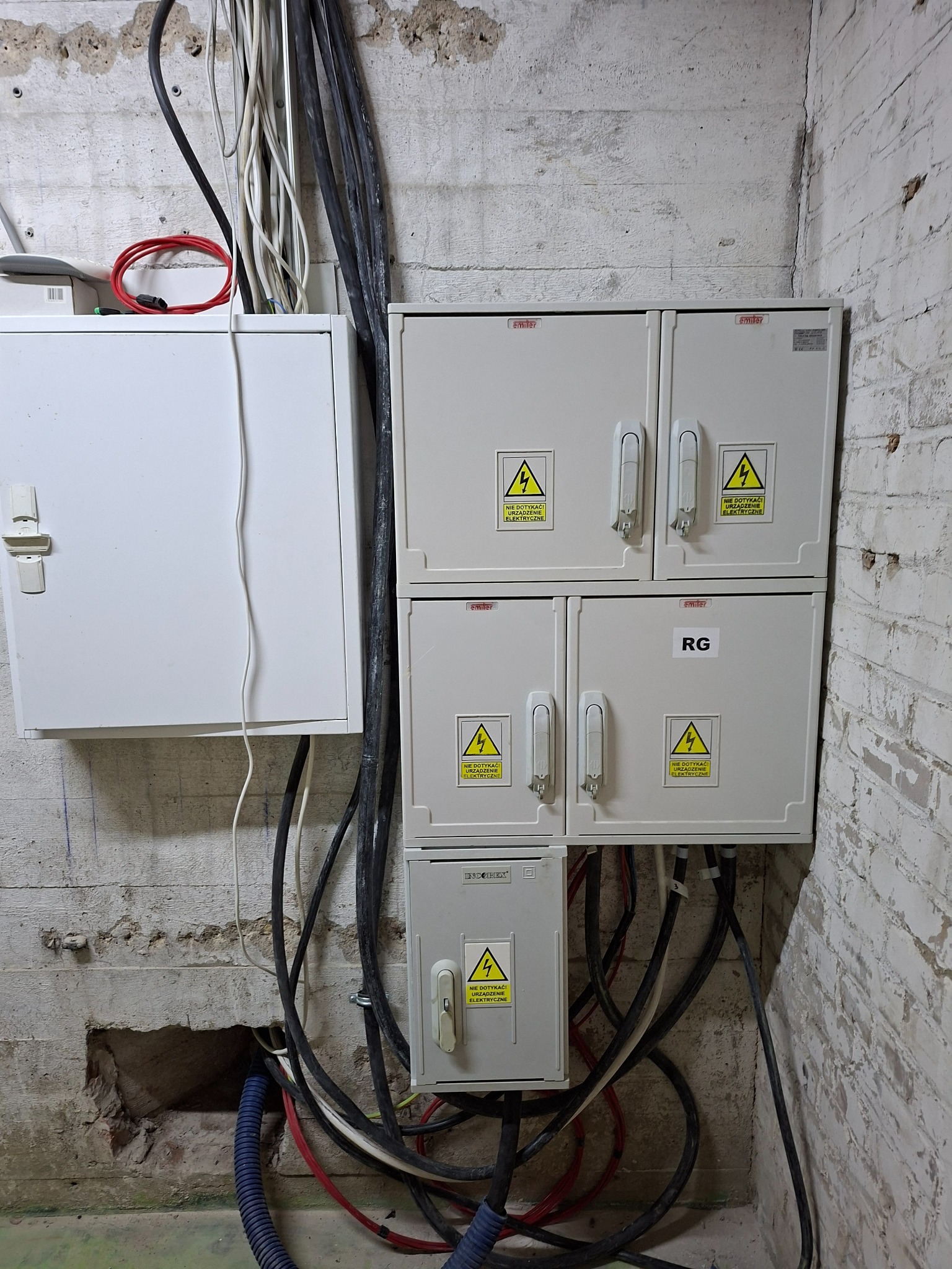 Sześć szafek elektrycznych, w tym cztery białe ułożone w stos, zamontowane na surowej betonowej ścianie. Widoczne liczne kable i ostrzeżenia.