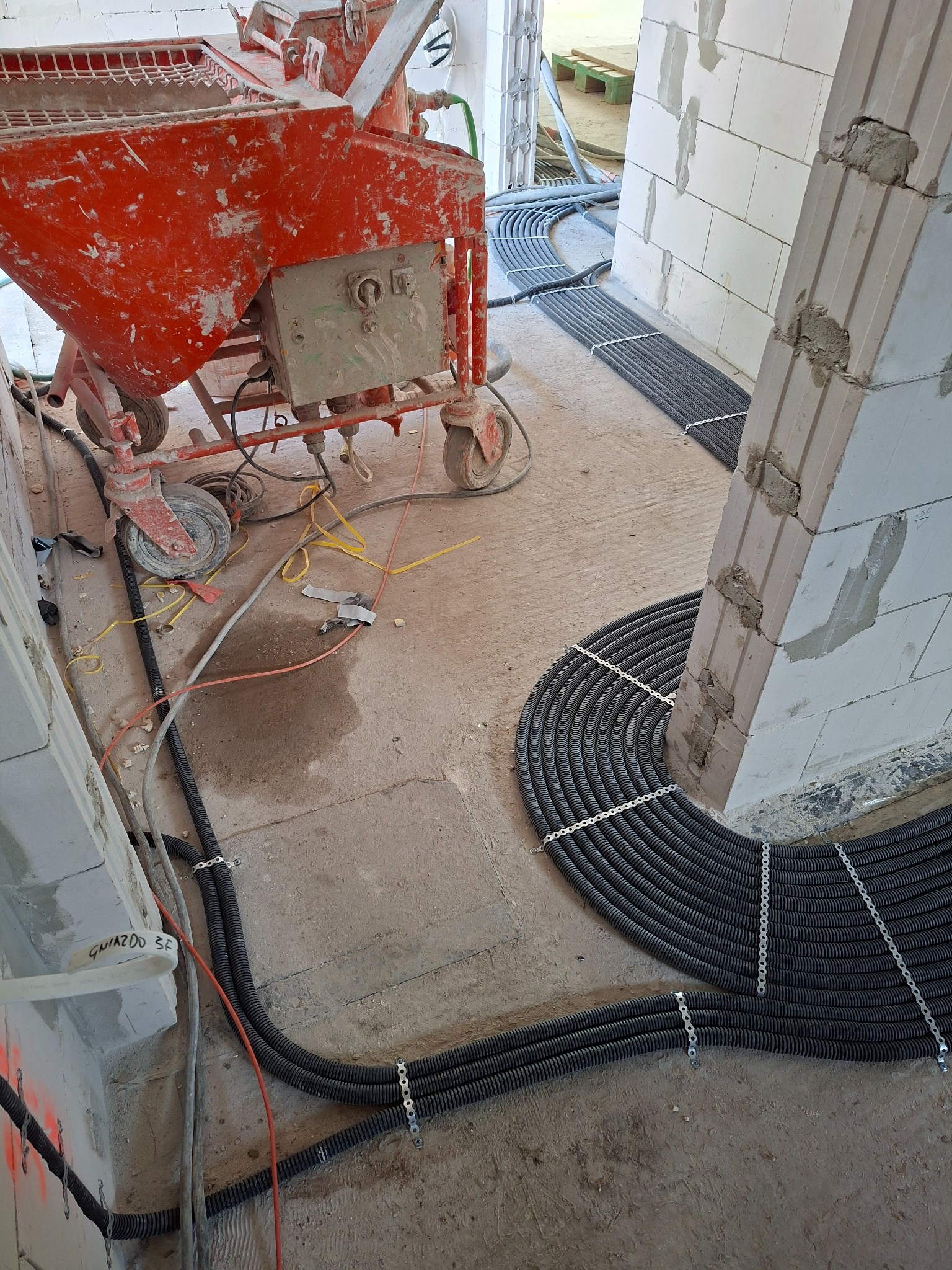 Czarne rury karbowane do instalacji elektrycznej, w tym oznaczone 'GNIAZDO 3F', starannie ułożone i przymocowane do betonowej posadzki na budowie, tworzące łuk wokół filaru. W tle czerwona maszyna...