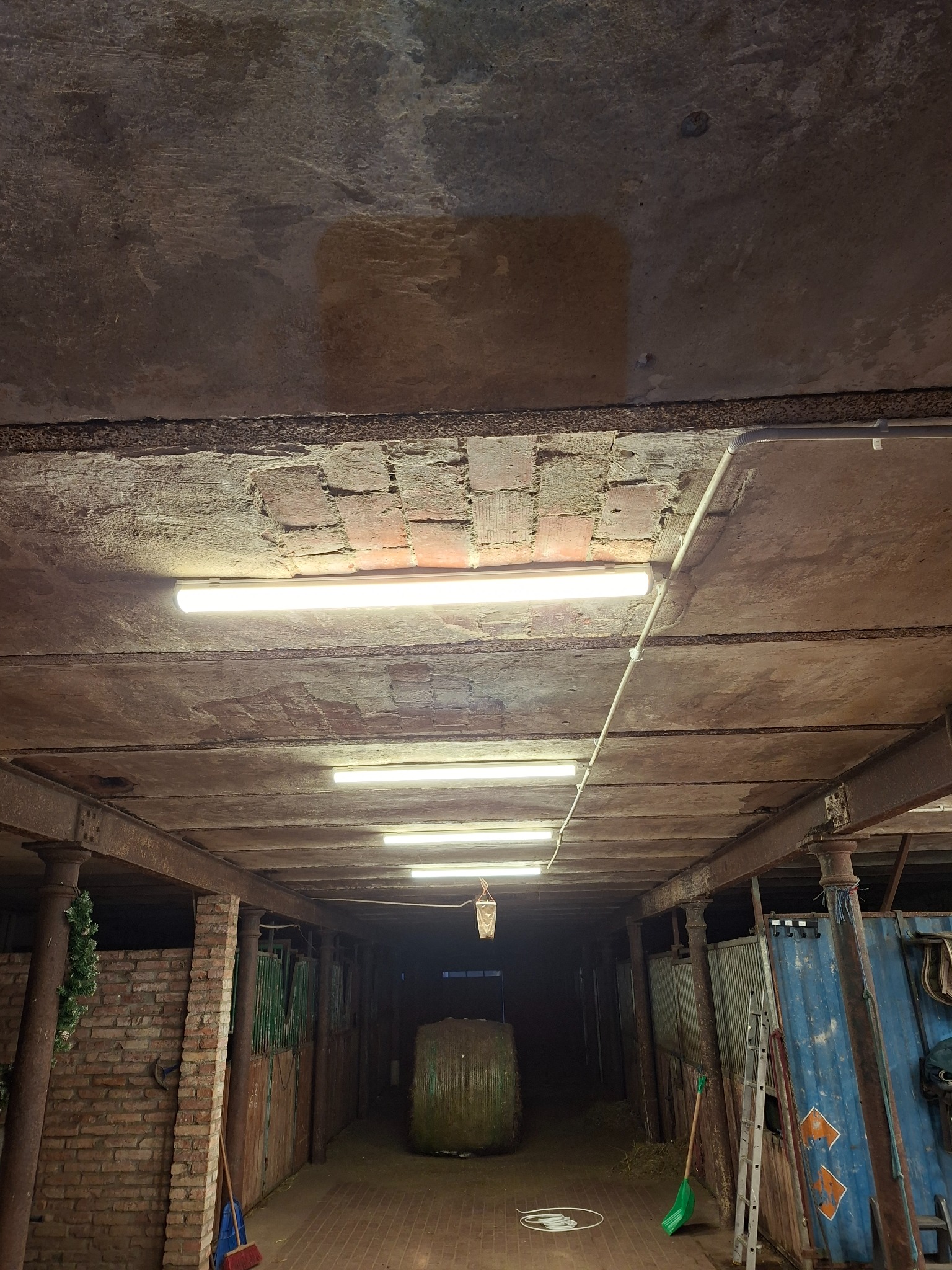 Oświetlony korytarz w starym budynku gospodarczym z rzędami podłużnych lamp LED zamontowanych na betonowym suficie, z widocznymi przewodami instalacyjnymi i metalowymi belkami.