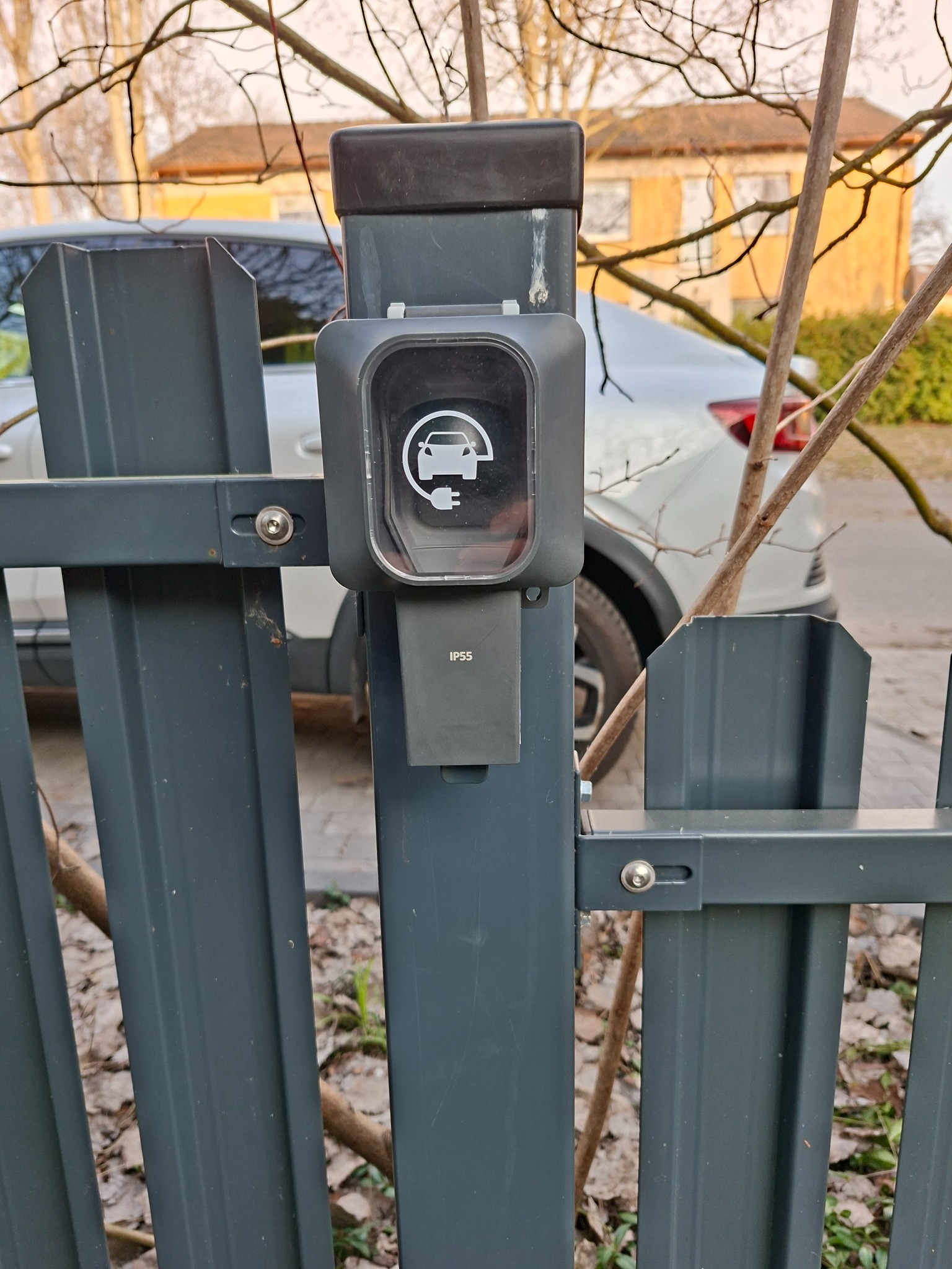 Szara stacja ładowania samochodów elektrycznych z ikoną auta i wtyczki, o klasie szczelności IP55, zamontowana na ciemnoszarym metalowym ogrodzeniu, z białym autem w tle.