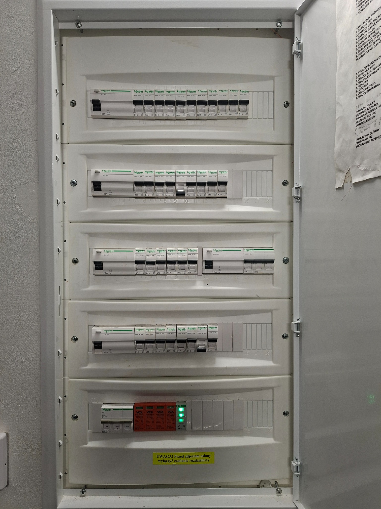 Otwarta biała rozdzielnica elektryczna z pięcioma rzędami wyłączników nadprądowych Schneider Electric, z zielonymi diodami na dole, żółtą naklejką ostrzegawczą i listą obwodów na drzwiach.
