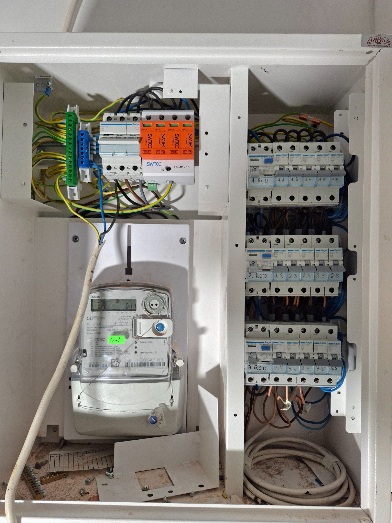 Otwarta rozdzielnica elektryczna z licznikiem ISKRA, modułami Hager i ogranicznikiem przepięć Simtec, ukazująca precyzyjne okablowanie.