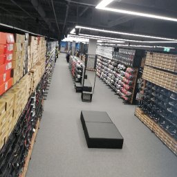 Sviatoslav Berezovskyi - Długi korytarz sklepu sportowego z rzędami półek wypełnionych pudełkami z obuwiem i odzieżą, oświetlony rzędami lamp sufitowych.