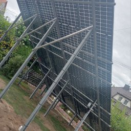 Sviatoslav Berezovskyi - Widok od tyłu na panele fotowoltaiczne zamontowane na metalowej konstrukcji na działce, z widoczną instalacją i elementami mocującymi, w tle dom jednorodzinny i zieleń.