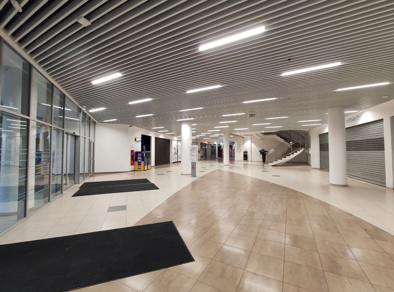 Przestronne, jasne wnętrze pasażu handlowego z widocznymi instalacjami oświetleniowymi pod sufitem, zamkniętymi roletami sklepów, szklanymi drzwiami wejściowymi i schodami spiralnymi w oddali.