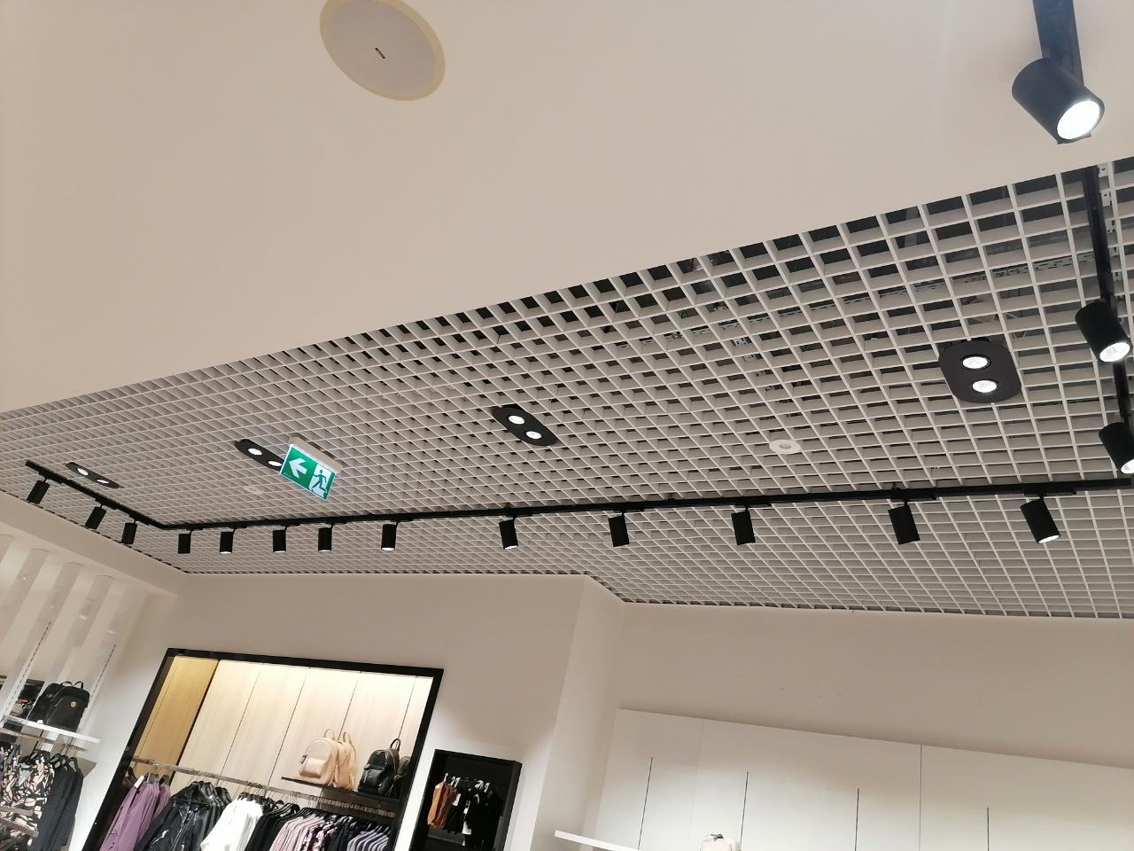 Sufit rastrowy z zainstalowanym systemem szynoprzewodów oświetleniowych, czarne reflektory punktowe, okrągły głośnik sufitowy i zielony znak ewakuacyjny w sklepie odzieżowym.