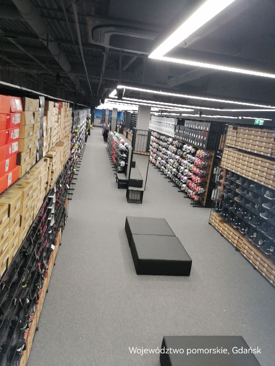 Długi korytarz sklepu sportowego z rzędami półek wypełnionych pudełkami z obuwiem i odzieżą, oświetlony rzędami lamp sufitowych.